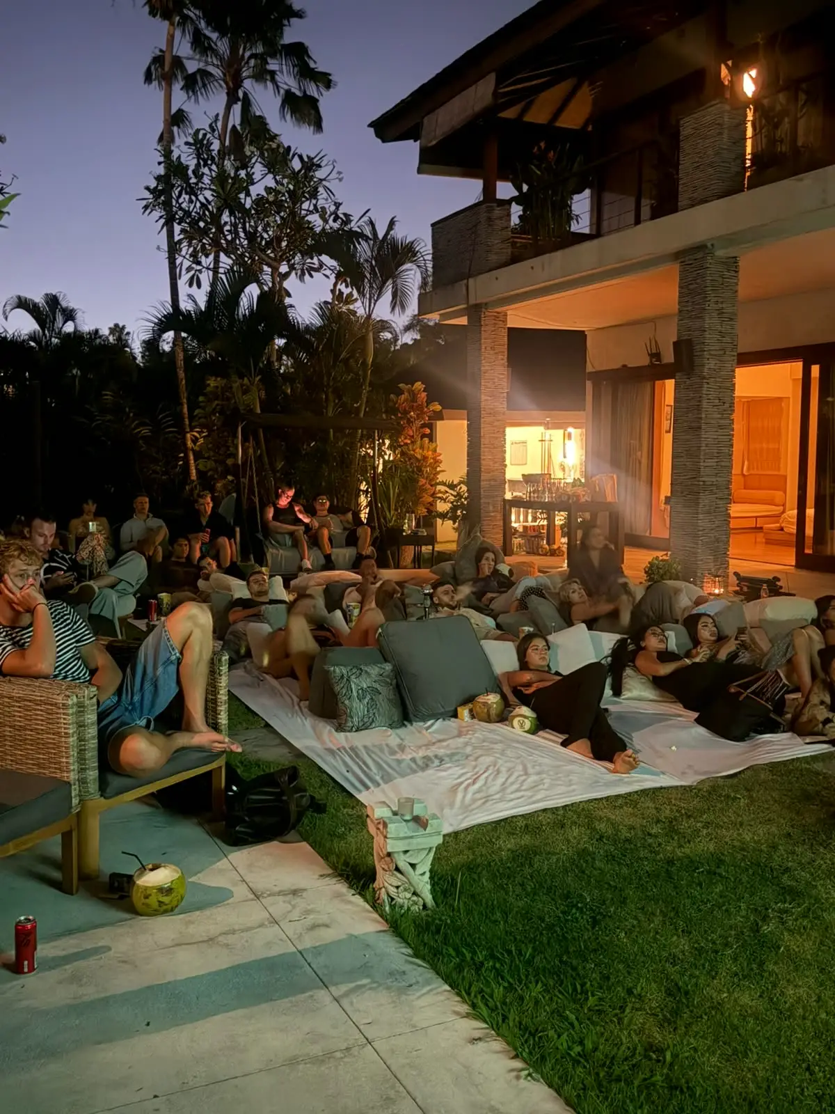 Guests on cushions on the lawn at dusk, the villa lit behind them