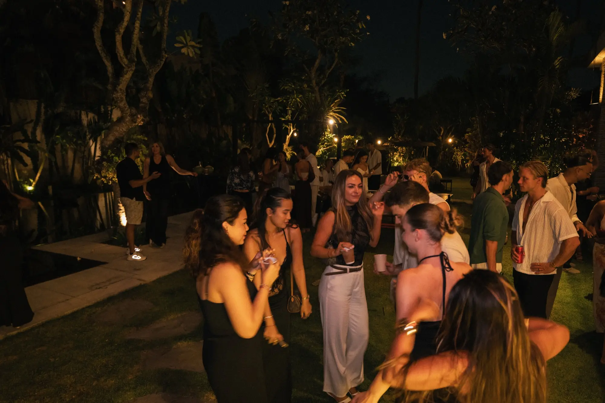 Guests talking on the garden lawn at night, the trees lit from below