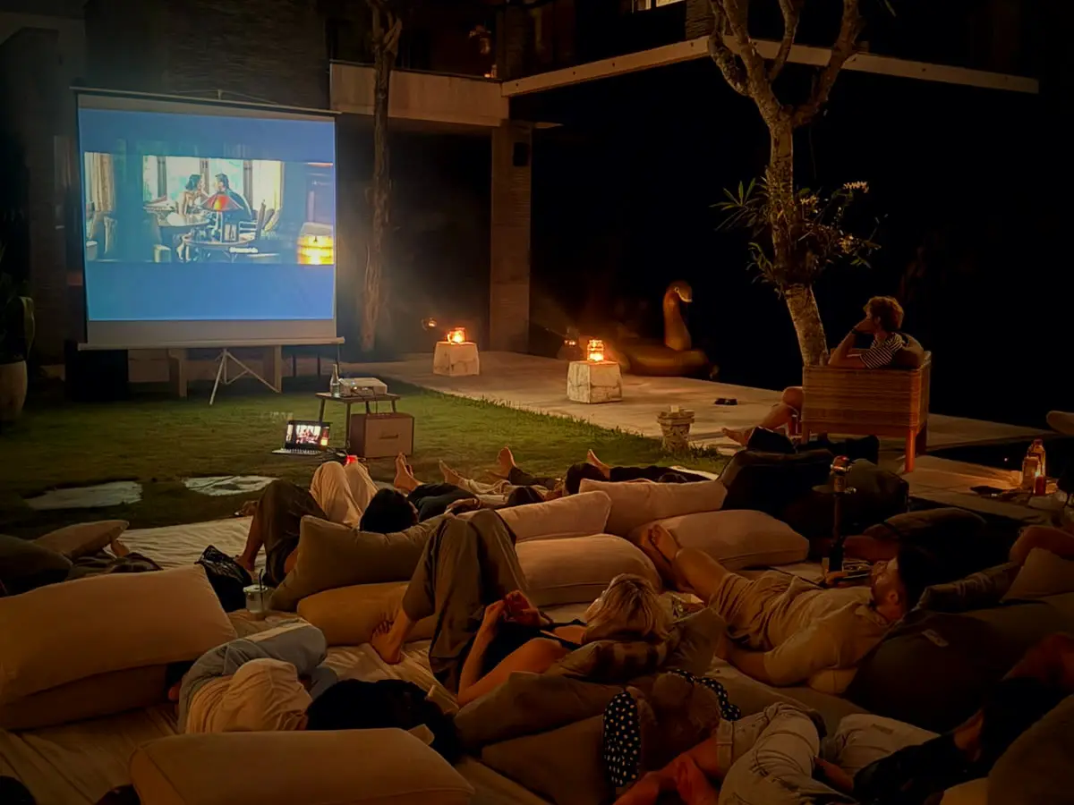 Guests lying on floor cushions in the villa garden at night, watching a film on the big screen by candlelight